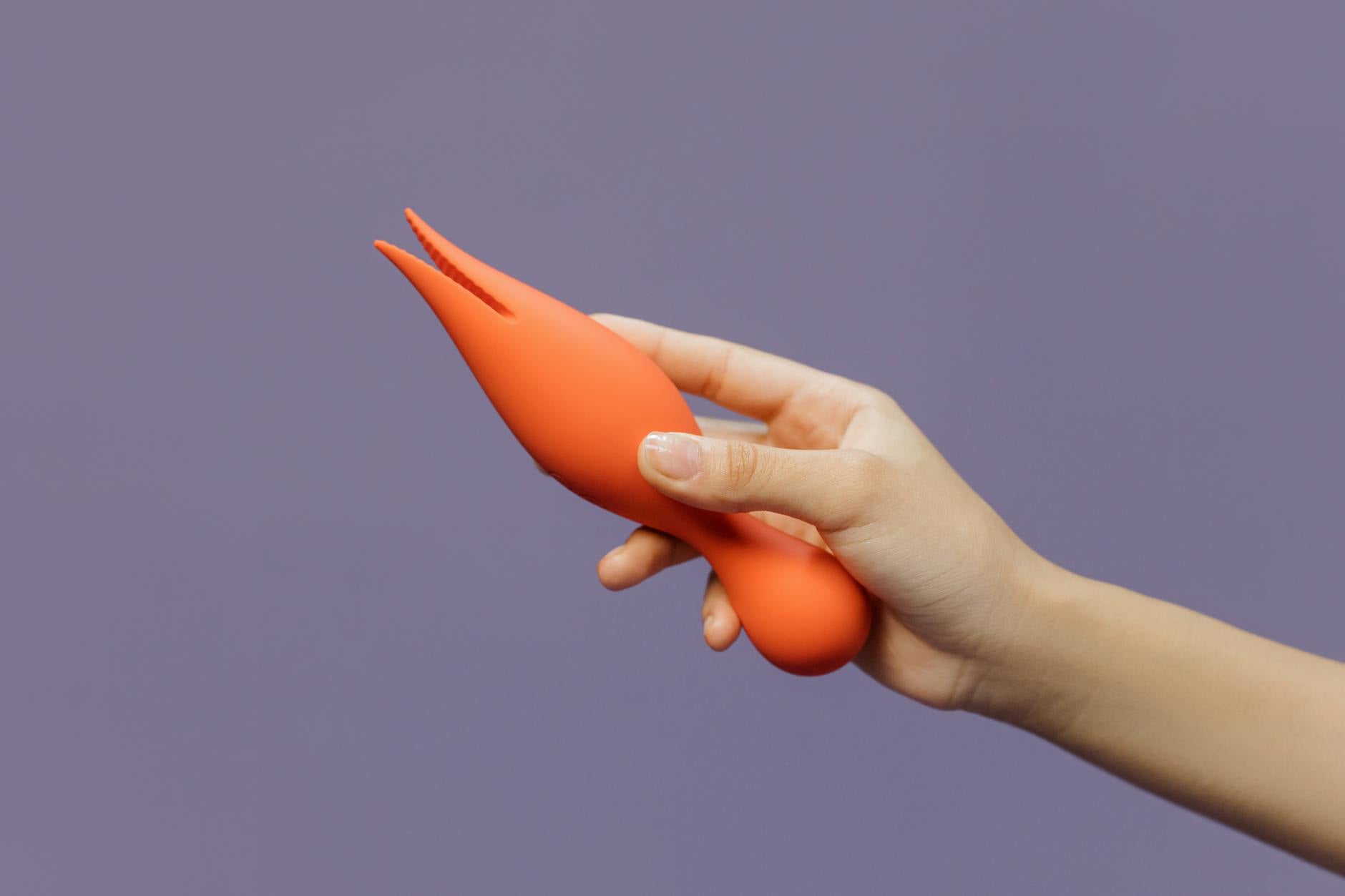 Eine Hand hält einen orangefarbenen Clitoralen Vibrator gegen einen minimalistischen violetten Hintergrund und zeigt moderne Sinnlichkeit.